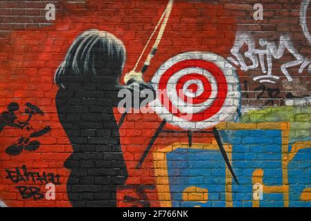 A female archer in a dark suit prepares to fire an arrow at a target on a brick wall sprayed bright red in psychedelic street art on one of the former long-term squatters’ strongholds close to the world-famous Slangenhuis or Snake House in Spuistraat, Amsterdam, North Holland, Netherlands. Stock Photo