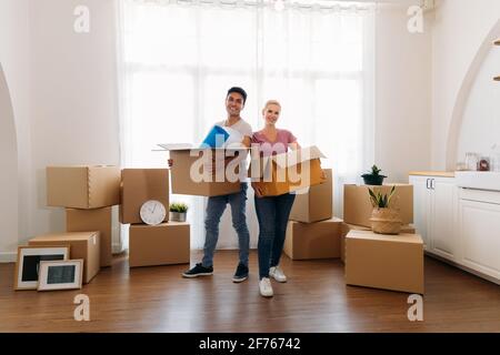 Portrait of happy young caucasian husband and wife posing in their new house and holding cardboard boxes while moving in and looking at camera Stock Photo