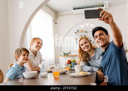 Caucasian Family group of four taking selfie photo while eating a breakfast on dining table. Father, Mother, Daughter and Son enjoying happy time together with kitchen in background. Stock Photo