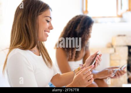 Two happy mixed race women friends having fun at home with social network chatting on smartphone with internet mobile connection technology. New norma Stock Photo