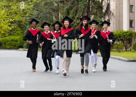 Cheerful multiracial students in graduation costumes walking by university campus Stock Photo