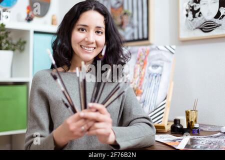 Young woman artist in her studio smiling showing different types of brushes. Selective focus. Concept of online learning.