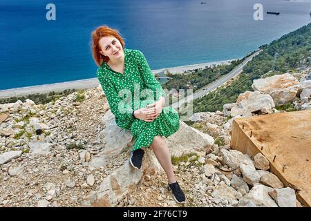 Smiling young European woman with red hair dressed in green summer dress sits on rock above deserted beach in Antalya near Mediterranean Sea. Stock Photo