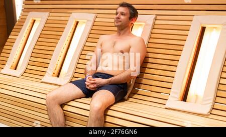 A beefy adult man warms his back on a wooden slatted bench with infrared heating lamps in a spa wellness center. Handsome man treating back pain in in Stock Photo