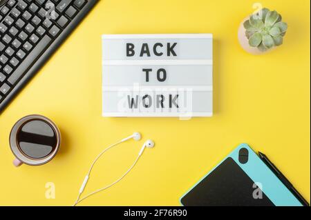 Back to work - text on display lightbox on yellow background workplace. Black keyboard, white earphones, coffee, tablet. Stock Photo