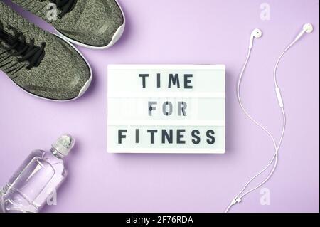 Time for fitness - motivanional text on display lightbox on purple background. Flat lay - Sneakers, a bottle of water, white headphones. Sport concept Stock Photo