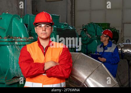 Young Worker in Red Coveralls and Hardhat Standing in Industrial Interior With Pipes and Generators. Portrait of Confident Young Machine Operator. Stock Photo