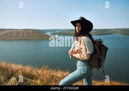 Adorable woman wearing casual clothing and cowboy hat standing on peak of mountain and looking at camera. Female traveler with backpack enjoying scenic view of Dniester canyon. Stock Photo