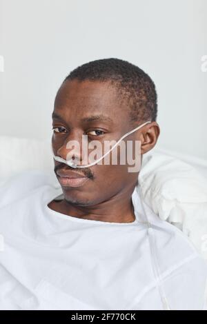 Vertical closeup portrait of African-American man lying in hospital bed with oxygen support tubes and looking at camera Stock Photo