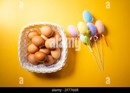 Basket with fresh eggs and fluffy colorful decorative plastic eggs isolated on yellow. Easter concept. Stock Photo