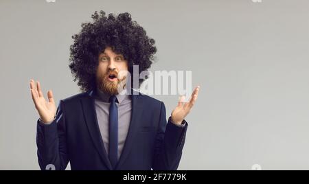 Funny young man in curly wig gasps in surprise standing on gray copy space background Stock Photo