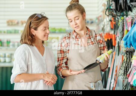 Happy young assistant of gardening shop in apron and shirt showing small shovels to mature brunette female customer in large supermarket Stock Photo