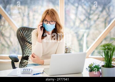 Portrait shot of middle aged woman wearing face mask and talking on mobile phone while working at the office during coronavirus pandemic. Stock Photo