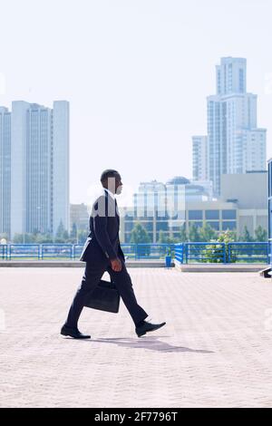 Young elegant African businessman in formalwear moving down sidewalk in front of camera against cityscape with skyscrapers and park Stock Photo