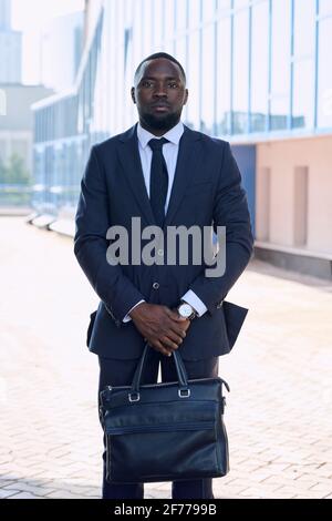Young confident African businessman in formalwear standing in front of camera against exterior of modern building in urban environment Stock Photo