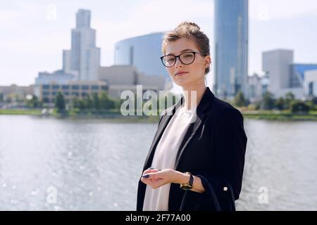 Young confident businesswoman in smart casualwear and eyeglasses looking at you while standing by riverside against cityscape Stock Photo