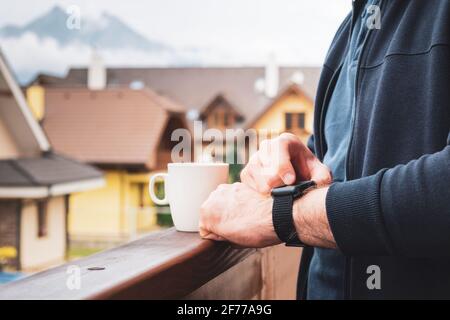Man using his smart watch on the balcony and drinking coffee with an amazing view on the mountains and roofs of houses Stock Photo