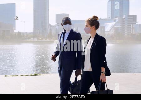 Young intercultural business professionals in suits and protective masks moving down sidewalk along riverside and discussing working plans Stock Photo