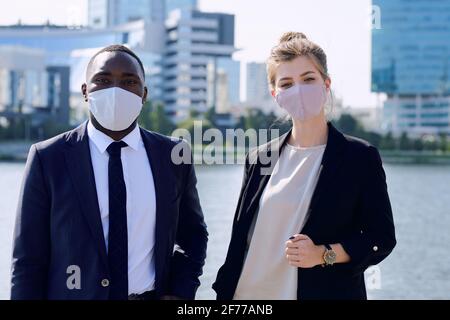 Two young intercultural managers in formalwear and protective masks looking at you while standing against riverside and skyscrapers Stock Photo
