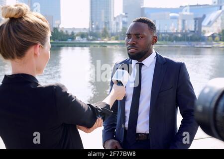 Young African entrepreneur standing in front of female reporter or journalist with microphone during interview in urban environment Stock Photo