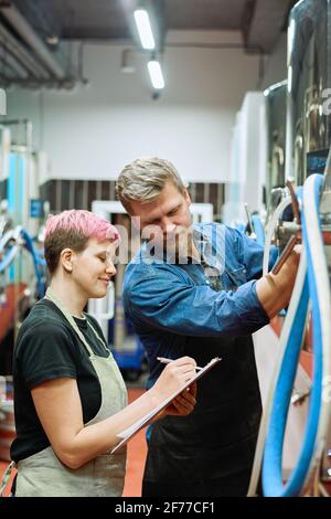 Happy young female worker of beer production factory making notes and talking to male colleague while both standing by processing machine Stock Photo