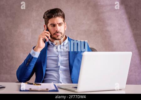 Shot of serious face businessman using his mobile phone and talking with somebody while sitting at office desk and woking on computer. Stock Photo