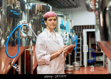 Young serious female brewery expert in whitecoat looking at you while using digital tablet against new equipment for beer production in plant Stock Photo