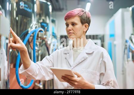 Young serious woman in whitecoat using digital tablet while standing in front of beer production equipment and touching control panel Stock Photo