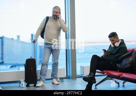 Mature man using smartphone during telephone conversation while young male traveler scrolling through contacts or posts in airport lounge Stock Photo