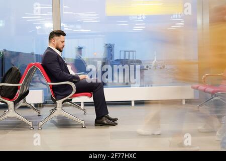 Side view of serious businessman in formalwear using laptop while sitting in lounge of modern airport and waiting for departure announcement Stock Photo