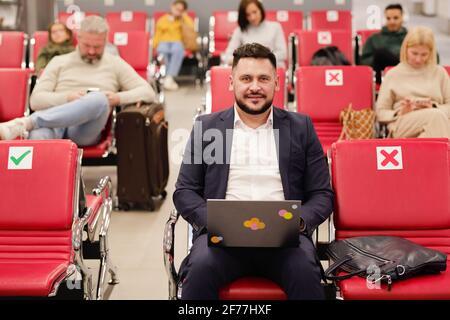 Smiling middle aged businessman in formalwear using laptop while sitting in lounge of modern airport against other people waiting for flight Stock Photo