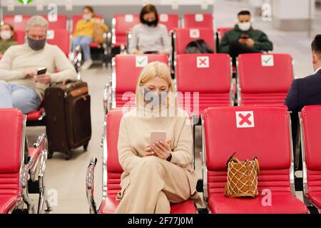 Mature blond female traveler in casualwear and protective mask using smartphone against other people waiting for flight announcement Stock Photo