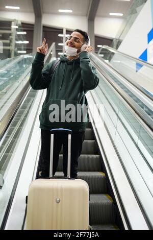 Contemporary happy young man in casualwear enjoying his favorite music in headphones while standing on step of moving escalator in airport Stock Photo