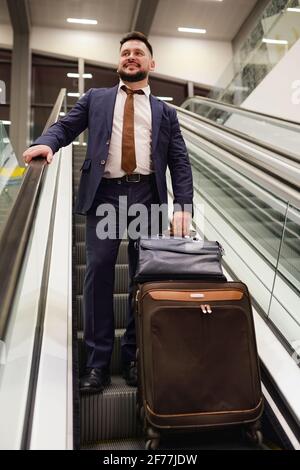 Cheerful and successful middle aged businessman in dark blue suit holding black leather handbag and suitcase while standing on step of escalator Stock Photo