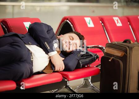 Tired middle aged businessman in formalwear lying on row of red leather seats and napping in lounge of modern airport while waiting for flight Stock Photo