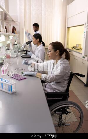Disabled scientist sitting at the table and working with samples in test tubes with her colleagues in the background Stock Photo