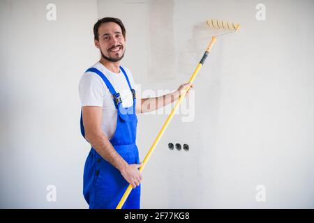 A bearded painter in work clothes paints the wall with a roller. He smiles friendly and poses for the camera Stock Photo