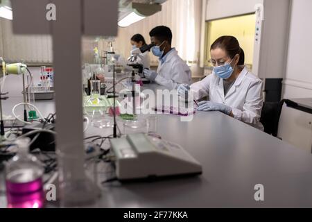 Young chemist in protective mask sitting at the table and working with chemical samples in the lab Stock Photo