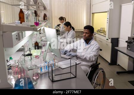 African disabled scientist sitting in wheelchair at the table and working on computer with his colleague in the background in the lab Stock Photo