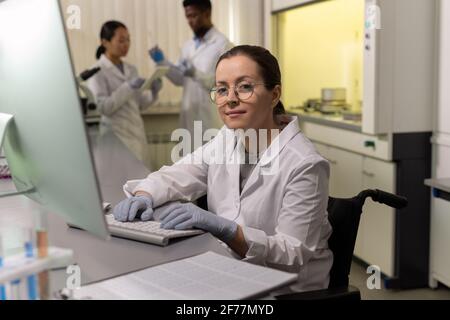 Portrait of female chemist in eyeglasses looking at camera while sitting at the table and working on computer in the laboratory Stock Photo