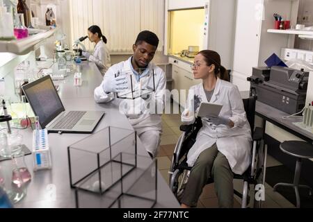 African young scientist holding mouse in the box with his colleague sitting in wheelchair and using digital tablet in the laboratory Stock Photo
