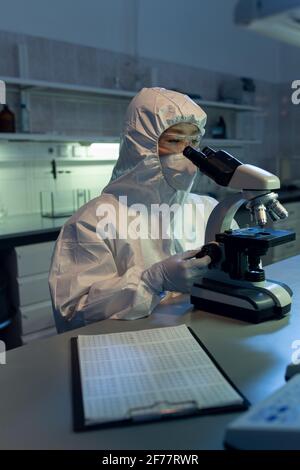 Female chemist in protective wear sitting at her workplace and looking through the microscope while working in the lab Stock Photo