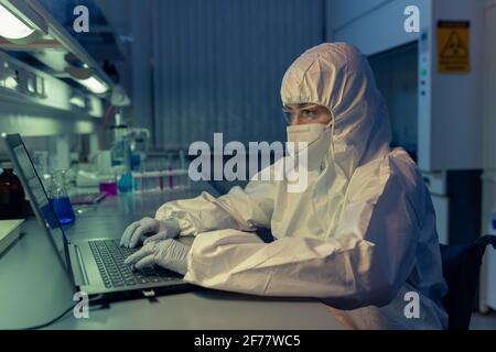 Scientist in protective workwear and mask typing on laptop at her workplace she working till late night in the lab Stock Photo