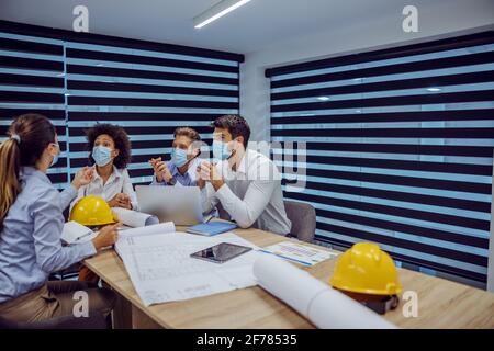 Multiracial group of architects with masks on faces sitting in boardroom, having meeting and discussing about project during corona outbreak. Stock Photo