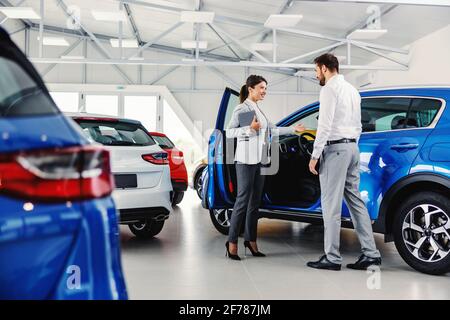 Friendly, smiling female seller showing brand new car to a customer while standing in car salon. Stock Photo