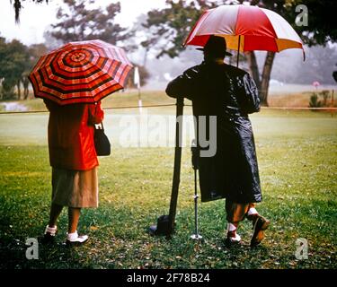 1970s COUPLE MIDDLE-AGED MAN AND WOMAN WEARING RAINCOATS CLEATED GOLF SHOES STANDING UNDER UMBRELLAS ON RAINING GOLF COURSE  - ks6369 HAR001 HARS SATISFACTION FEMALES MARRIED RURAL SPOUSE HUSBANDS HEALTHINESS COPY SPACE FULL-LENGTH LADIES RAINY PERSONS MALES UMBRELLAS GOLFING RAINING ATHLETIC SPECTATORS MIDDLE-AGED GALLERY PARTNER MIDDLE-AGED MAN SKILL ACTIVITY AMUSEMENT COURSE HAPPINESS MIDDLE-AGED WOMAN WELLNESS ADVENTURE GOLFERS HOBBY LEISURE INTEREST AND CHOICE HOBBIES KNOWLEDGE RECREATION PASTIME PLEASURE CONCEPTUAL DELAYED LINKS STYLISH SUPPORT RAINCOATS FANS RELAXATION WIVES AMATEUR Stock Photo