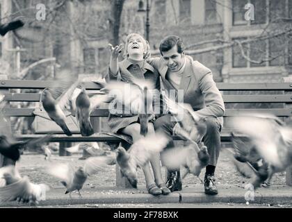 1970s HAPPY LAUGHINGCOUPLE IN URBAN PARK SITTING ON BENCH FEEDING PIGEONS  - r22903 HAR001 HARS OLD FASHION 1 FEEDING LAUGH YOUNG ADULT PLEASED JOY LIFESTYLE BIRDS FEMALES UNITED STATES COPY SPACE FRIENDSHIP FULL-LENGTH LADIES PERSONS UNITED STATES OF AMERICA MALES B&W DATING WING HAPPINESS CHEERFUL ADVENTURE DISCOVERY EXCITEMENT ATTRACTION NYC COURTSHIP MOTION BLUR CONCEPTUAL NEW YORK CITIES JOYFUL PIGEON VERTEBRATE WARM-BLOODED NEW YORK CITY POSSIBILITY FEATHERED PIGEONS SOCIAL ACTIVITY TOGETHERNESS WINGED YOUNG ADULT MAN YOUNG ADULT WOMAN BIPEDAL BLACK AND WHITE CAUCASIAN ETHNICITY COURTING Stock Photo