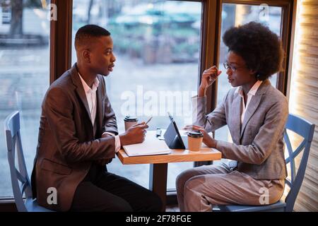 African business colleagues discussing working issues during meeting at modern cafe. Young man and woman sitting at table with modern gadgets and cups of coffee. Stock Photo