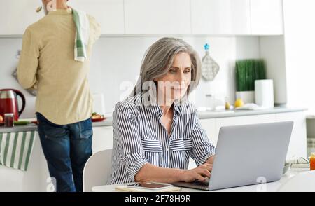 Mature wife remote working from home office while husband cooking in kitchen. Stock Photo