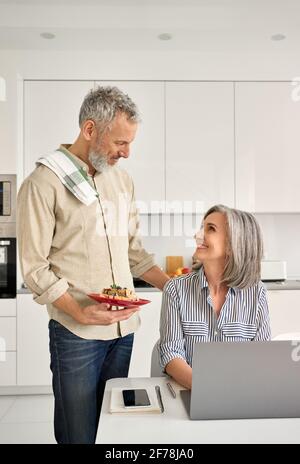 Happy mature wife working online at home office, husband cooking in kitchen. Stock Photo